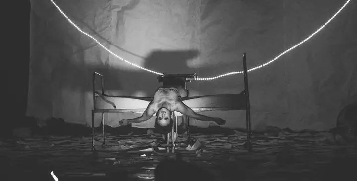 Escena en blanco y negro: una intérprete desnuda, tumbada boca arriba sobre una cama de hierro con la cabeza colgando hacia el suelo, bajo dos guirnaldas de bombillas encendidas.