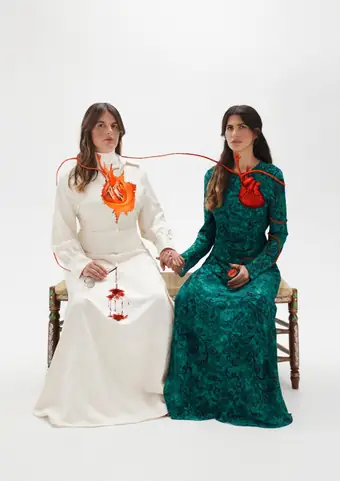 Cartel con dos mujeres sentadas de la mano sobre fondo blanco, unidas por un cordón rojo que sale de sus pechos.