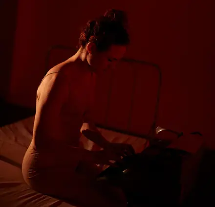 Escena bañada en luz roja: una mujer desnuda, sentada al borde de una cama de hierro, escribe en una máquina de escribir.