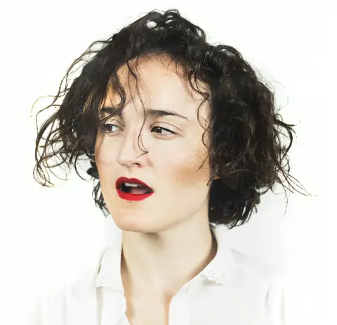 Retrato de estudio de Ángela Palacios sobre fondo blanco: pelo rizado corto, labios rojos y camisa blanca.