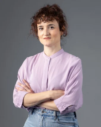 Retrato de Ángela Palacios con camisa lila y los brazos cruzados, sonriendo, sobre fondo gris.