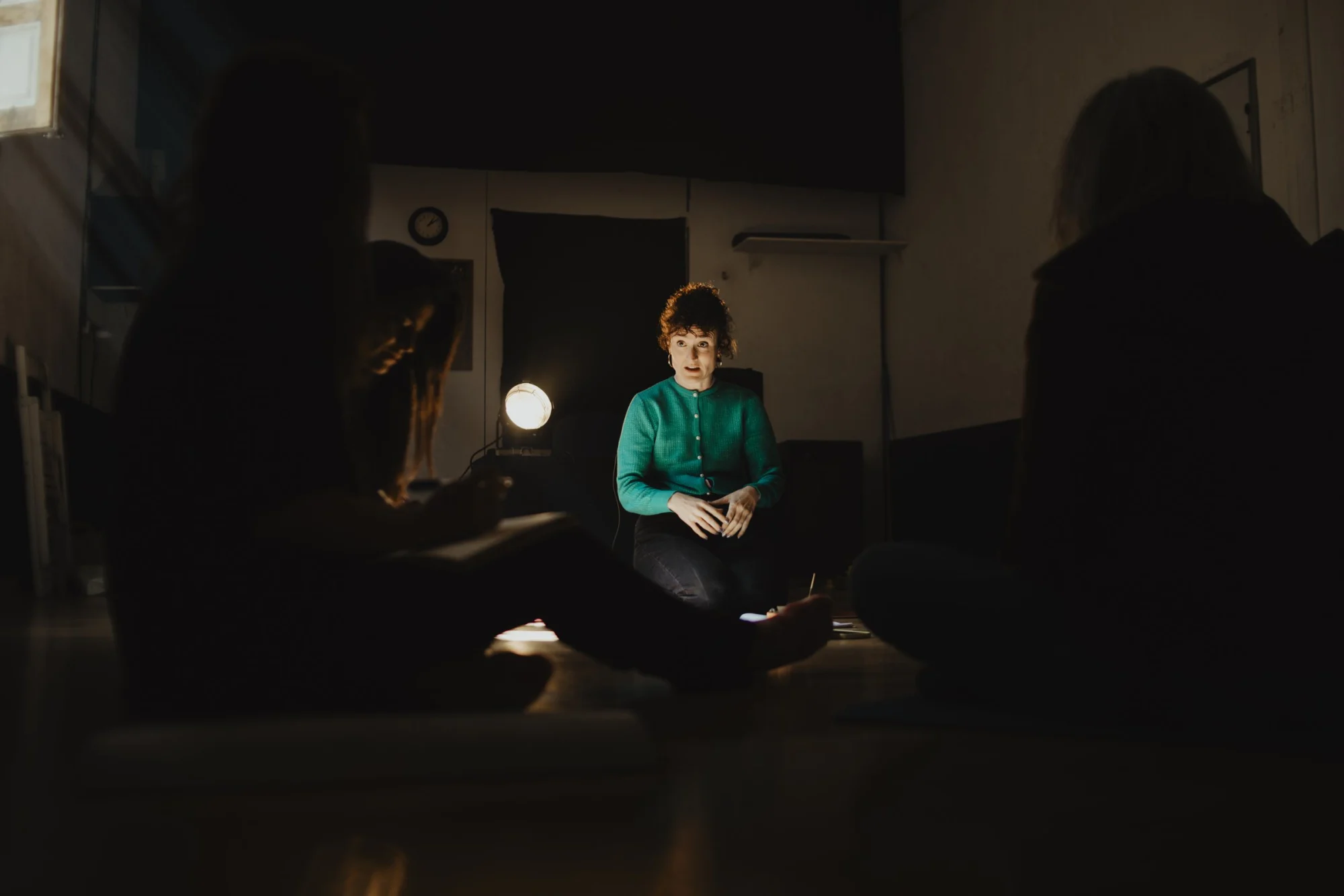 Ángela Palacios sentada en el suelo de una sala de ensayo en penumbra, rodeada de alumnas que la escuchan.