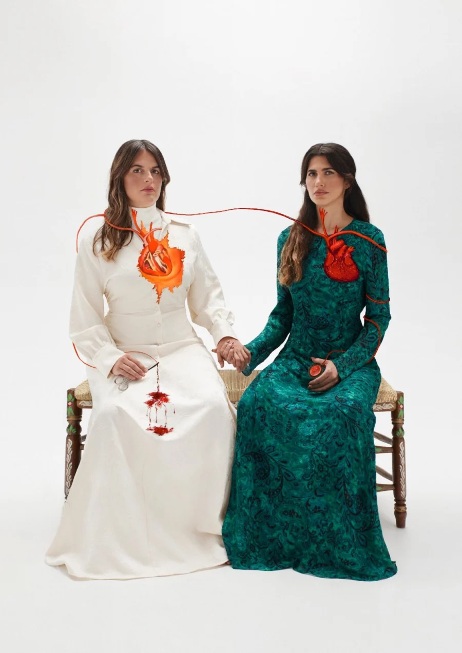 Cartel con dos mujeres sentadas de la mano sobre fondo blanco, unidas por un cordón rojo que sale de sus pechos.