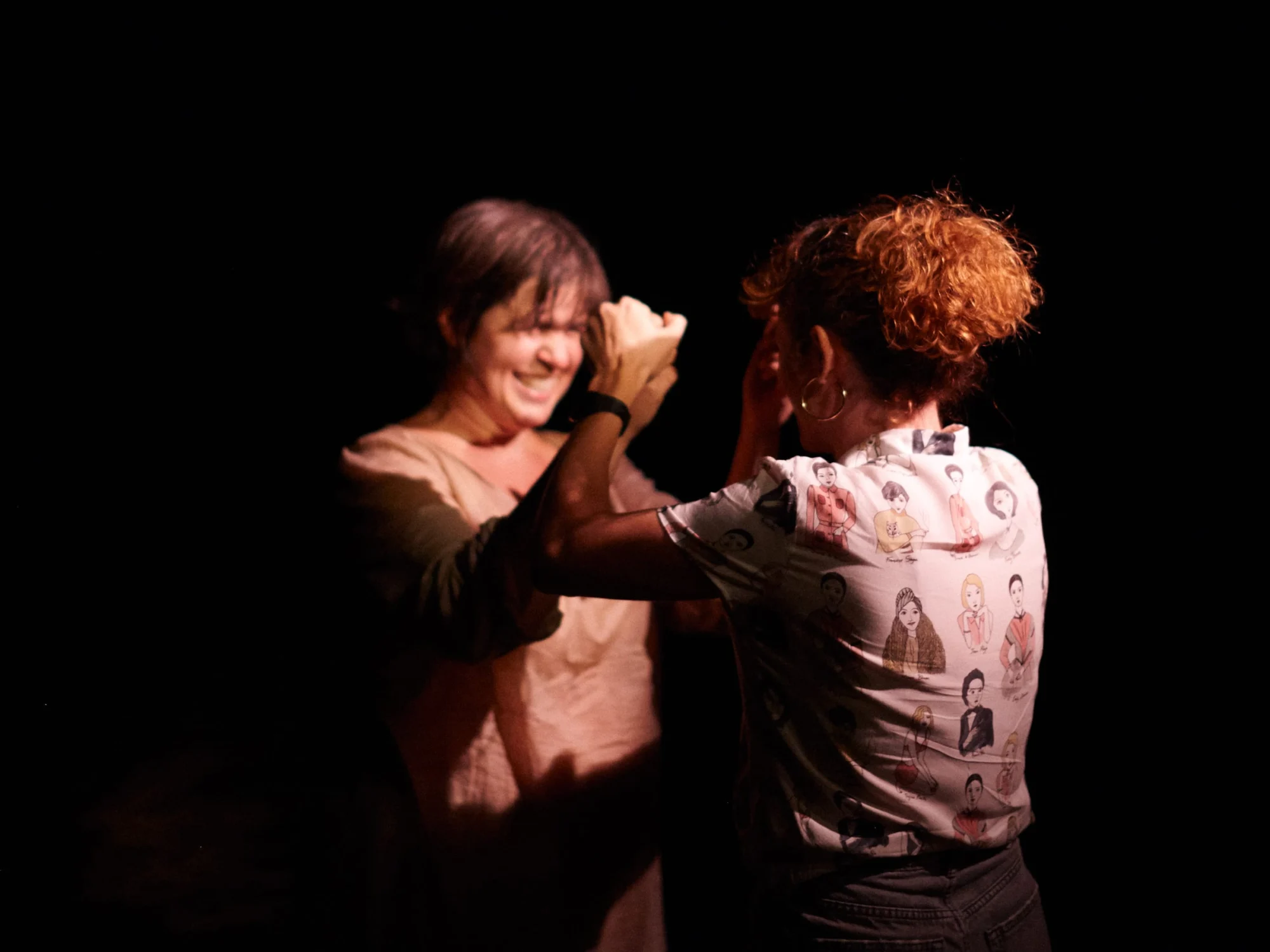 Escena de 200.000 dones: dos mujeres frente a frente, riendo, una con la mano junto a la cara de la otra, sobre fondo negro.