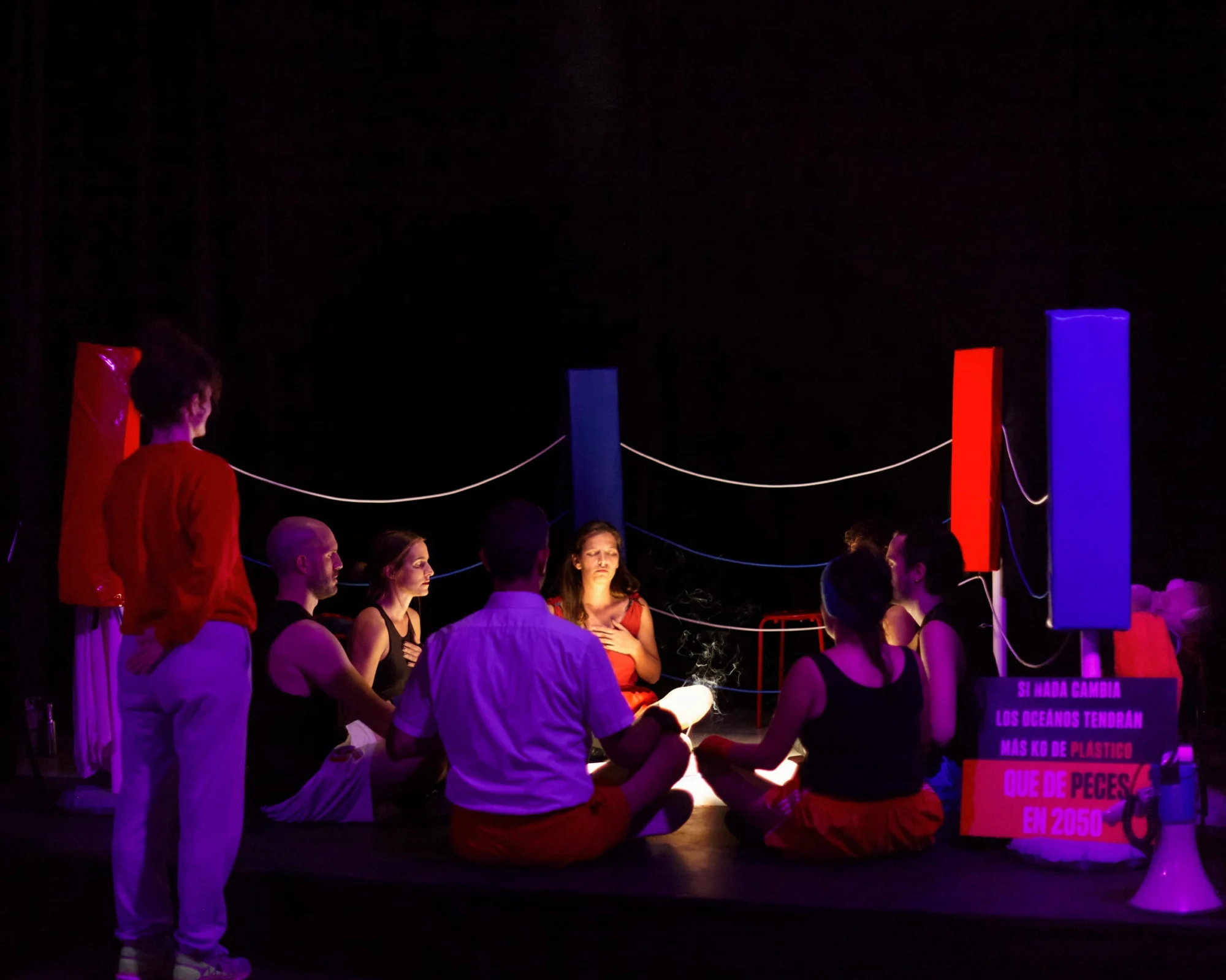 Un grupo de intérpretes sentados en círculo en un escenario en penumbra morada, entre columnas rojas y azules.