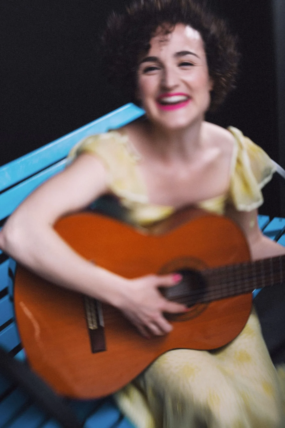 Ángela Palacios en Mirta en espera: vestido amarillo, sentada en un banco azul, tocando la guitarra y riendo.