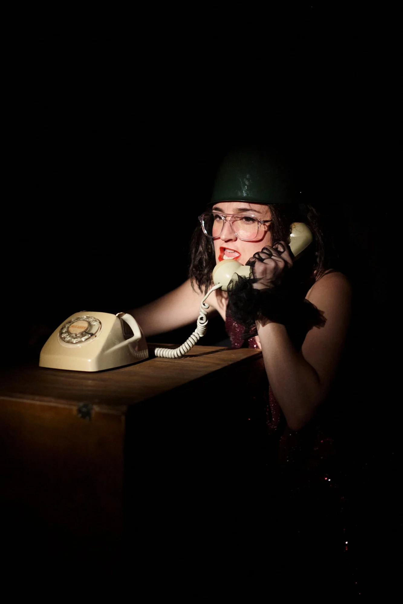 Ángela Palacios con casco y gafas, hablando por un teléfono de disco antiguo, en penumbra.