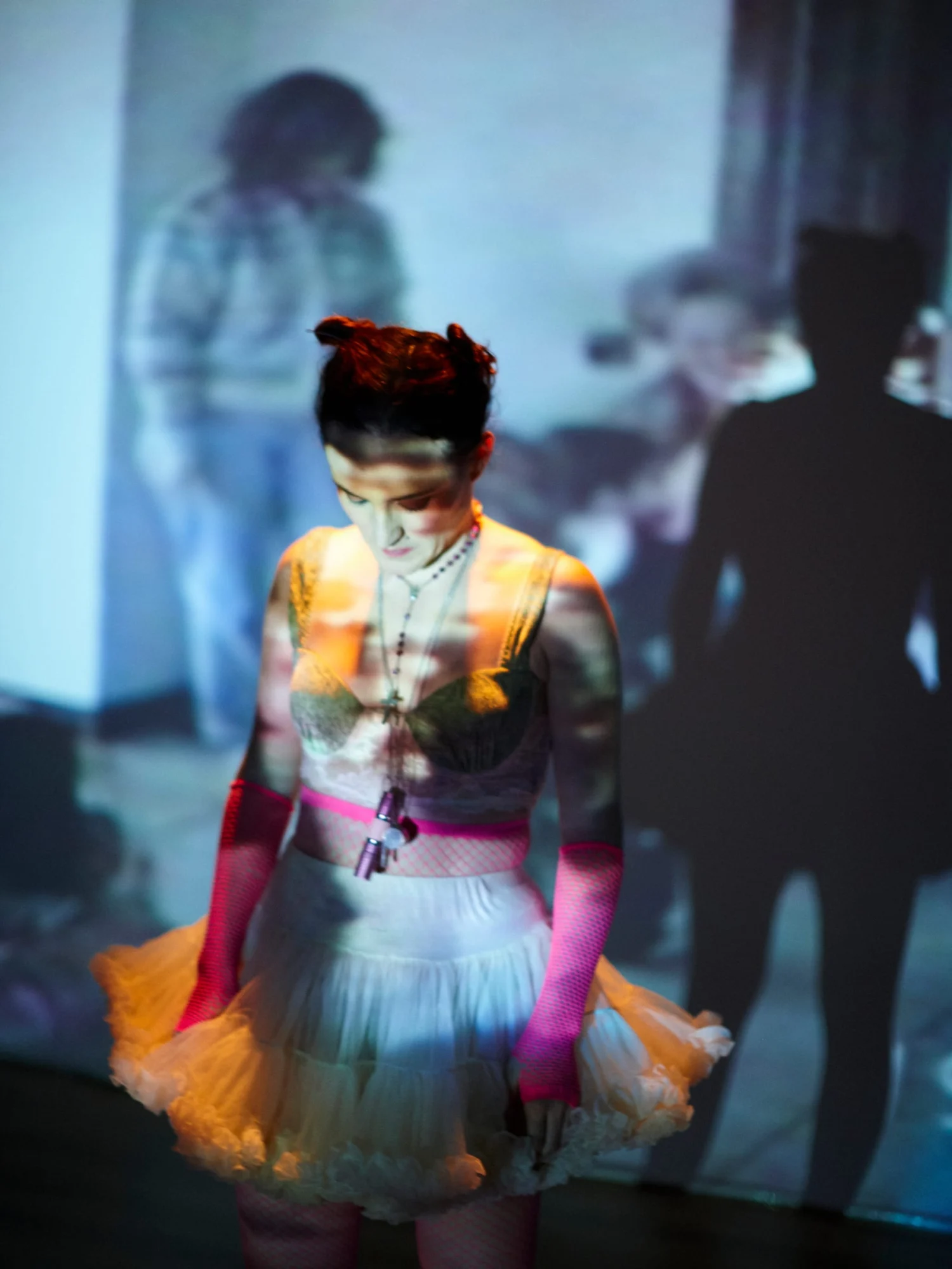 Ángela Palacios en escena, con tutú naranja, guantes de rejilla rosas y la mirada baja, bajo una proyección azulada con siluetas.
