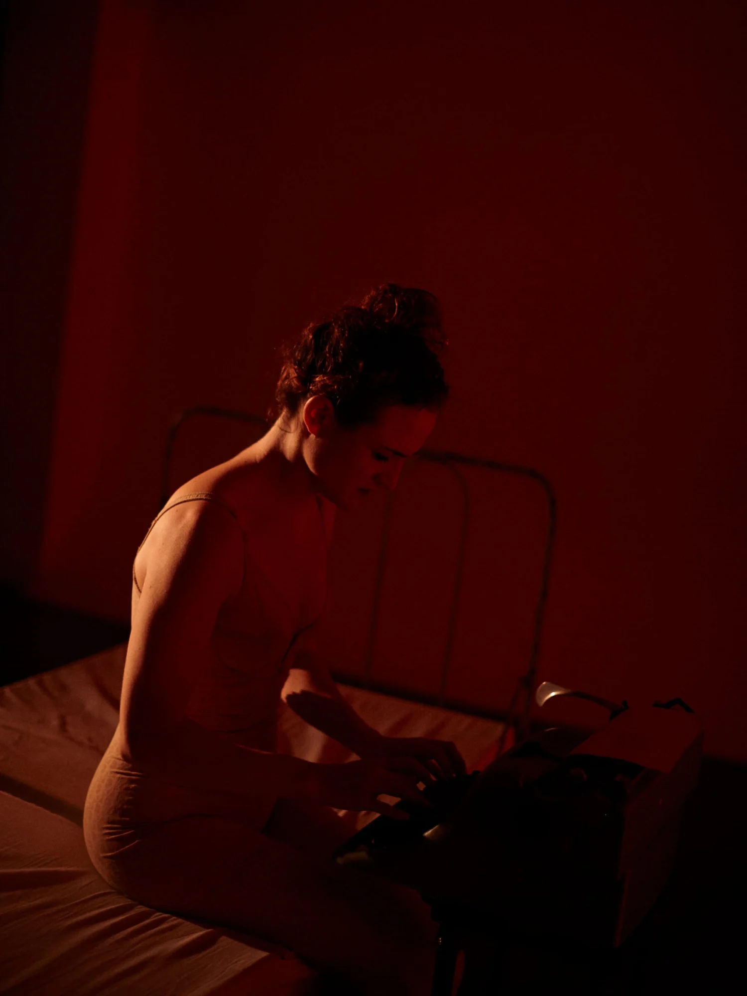 Una intérprete sentada al borde de una cama de hierro, inclinada sobre una máquina de escribir, en penumbra roja.