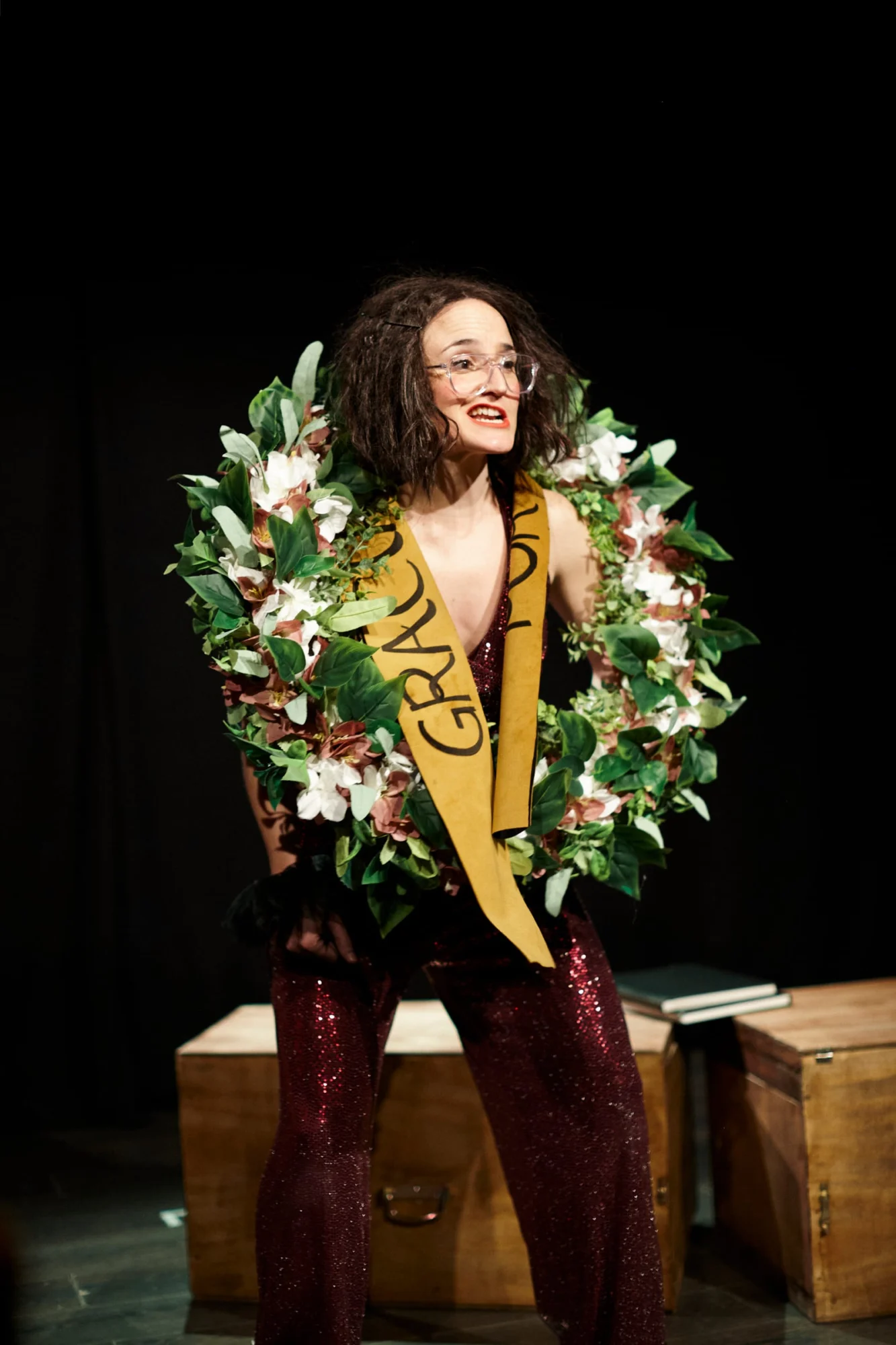 Ángela Palacios en escena, sonriendo, con una corona de flores al cuello y una banda amarilla cruzada, sobre fondo negro.