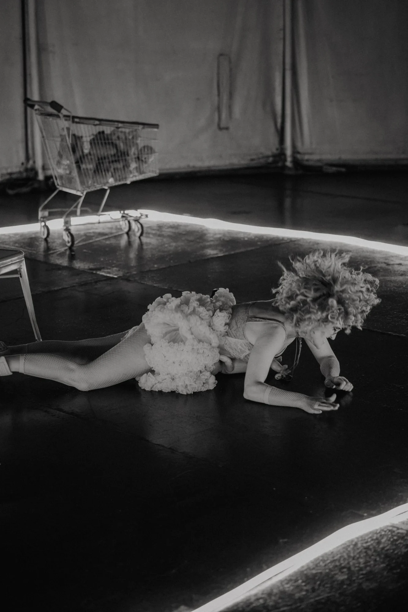 Escena en blanco y negro: una intérprete con tutú, tumbada boca abajo en el suelo del escenario junto a una línea de luz y un carro de la compra.