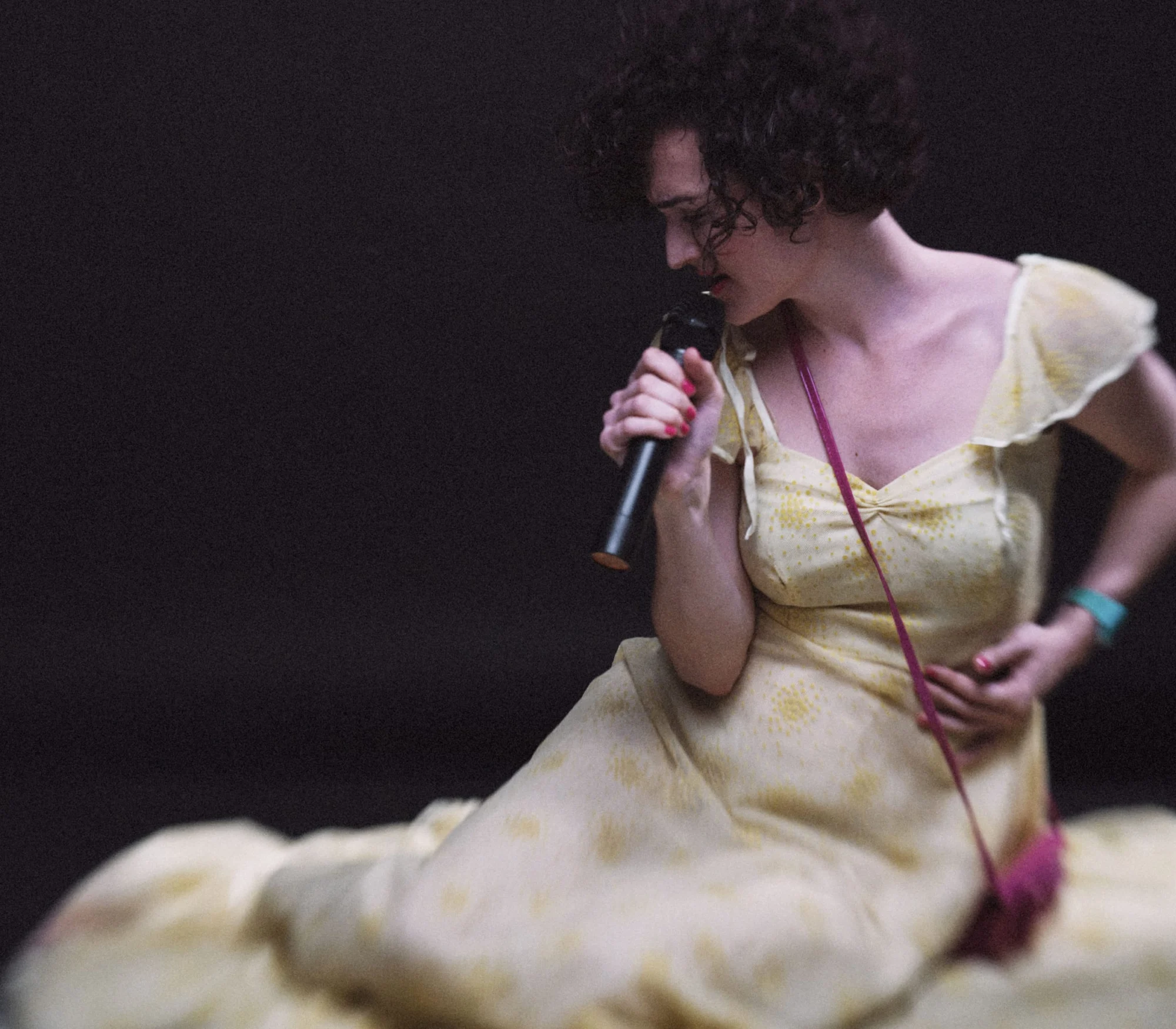 Ángela Palacios con vestido amarillo claro, sentada en el suelo y cantando a un micrófono sobre fondo oscuro.